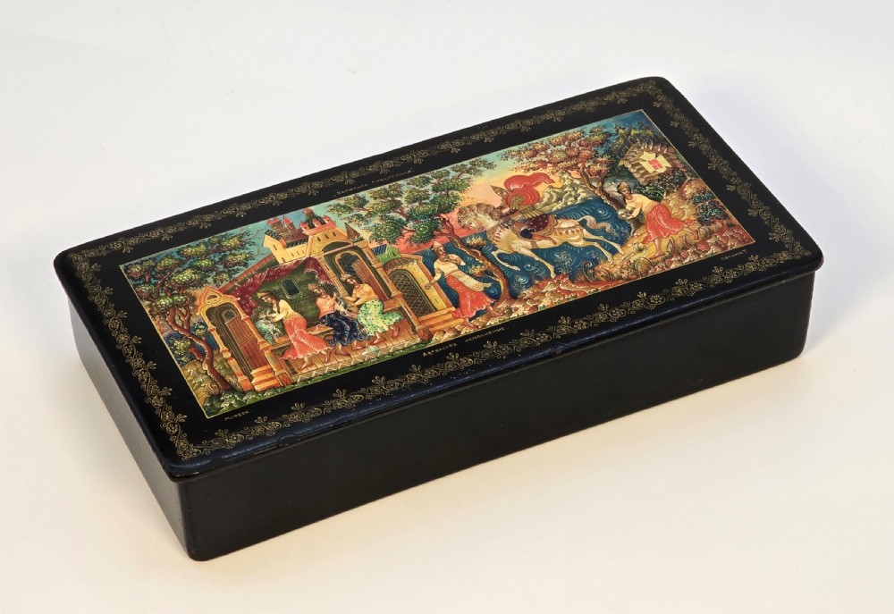 A rectangular black lacquered papier-mache box featuring a detailed hand-painted scene from Russian folklore framed by a delicate gold filigree border. View II
