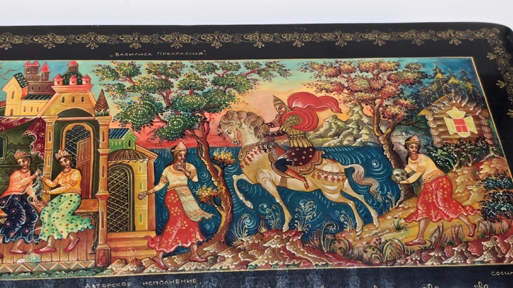A rectangular black lacquered papier-mache box featuring a detailed hand-painted scene from Russian folklore framed by a delicate gold filigree border. View IX