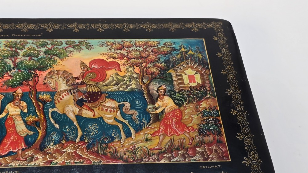 A rectangular black lacquered papier-mache box featuring a detailed hand-painted scene from Russian folklore framed by a delicate gold filigree border. View X