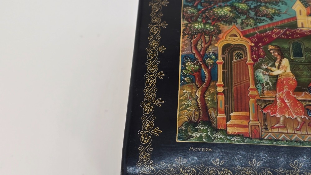 A rectangular black lacquered papier-mache box featuring a detailed hand-painted scene from Russian folklore framed by a delicate gold filigree border. View XII
