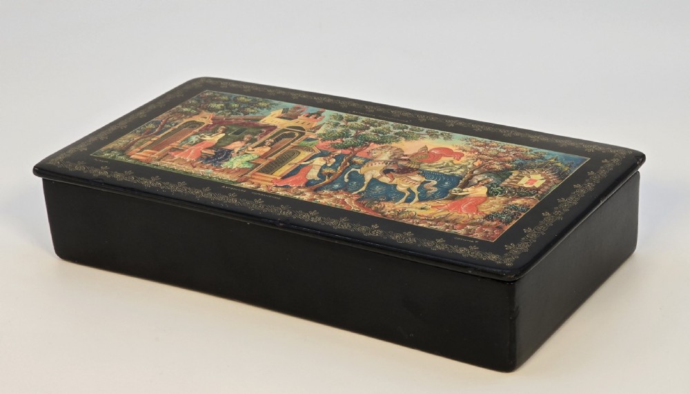 A rectangular black lacquered papier-mache box featuring a detailed hand-painted scene from Russian folklore framed by a delicate gold filigree border. View XIV