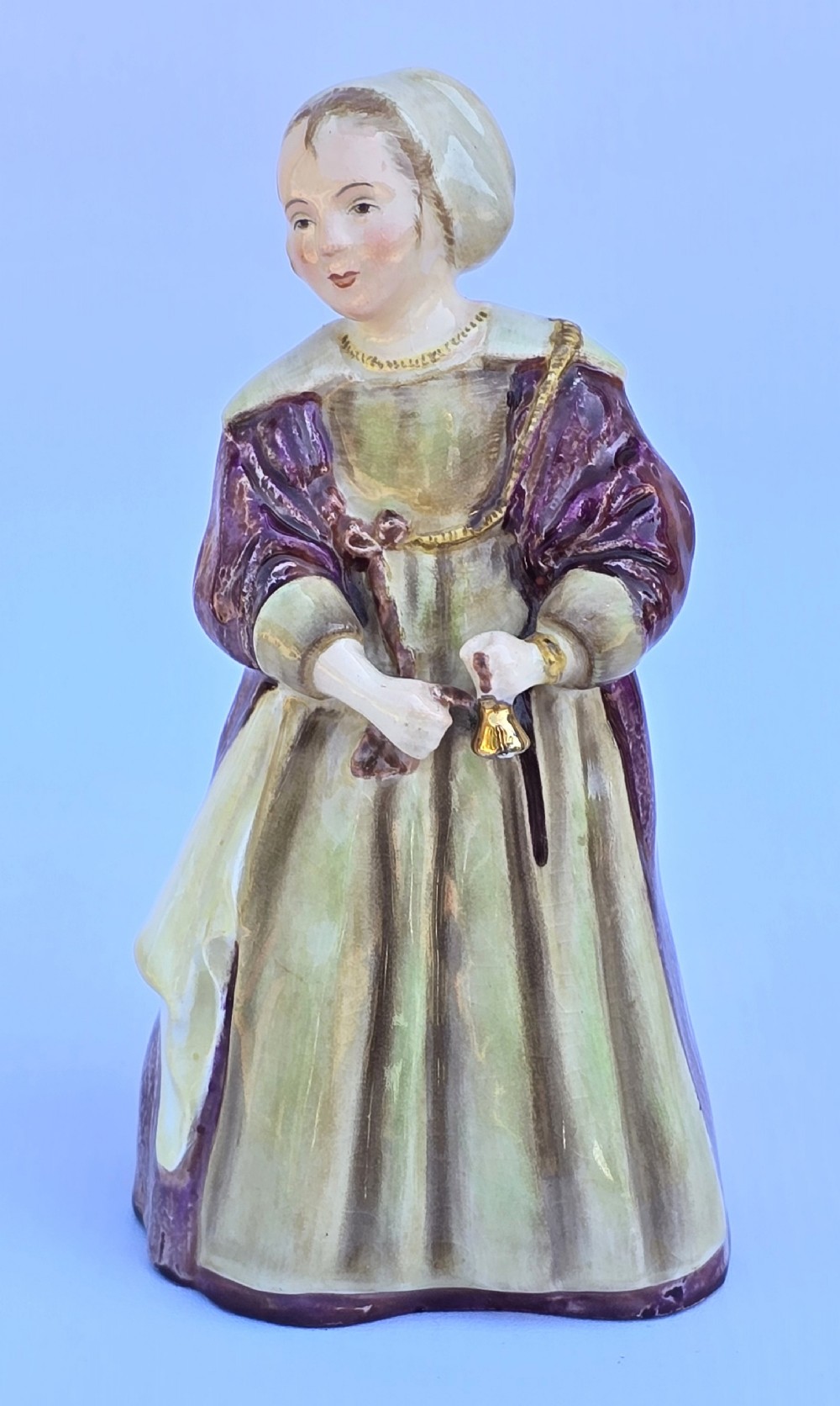 An Edwardian glazed china figurine depicting a young girl in a purple shawl and green dress holding a small gold bell.