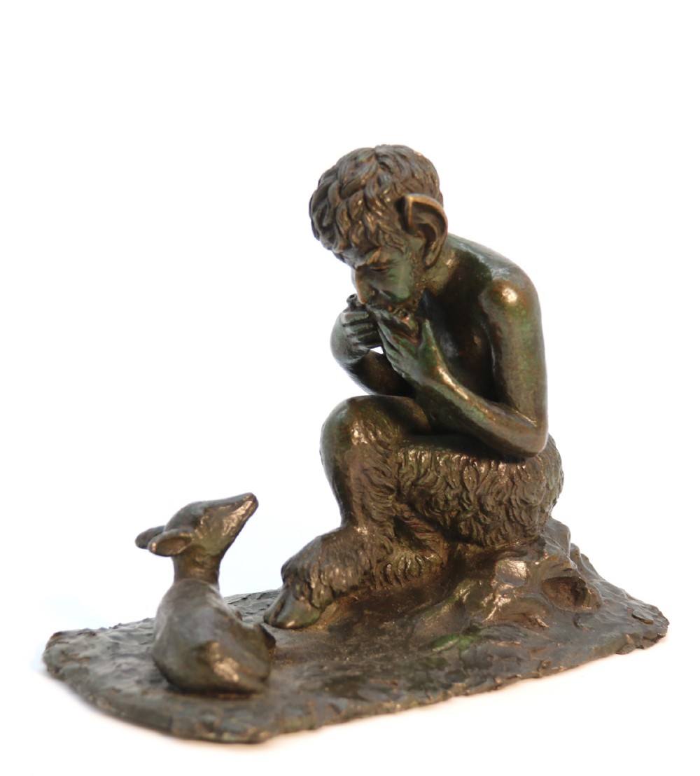 A dark brown patinated bronze sculpture depicting the mythological figure Pan crouching while playing panpipes to a small fawn sitting before him.
