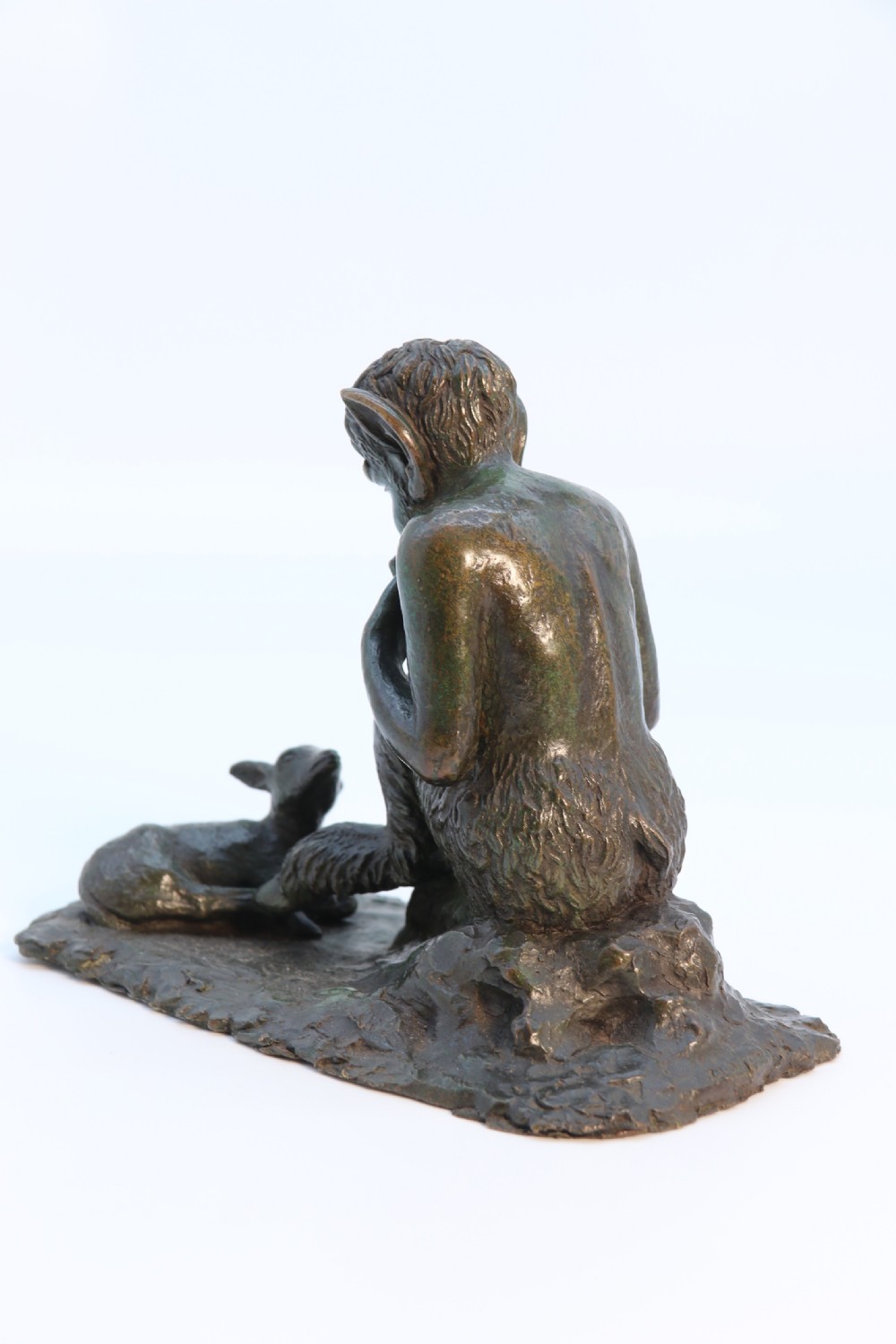 A dark brown patinated bronze sculpture depicting the mythological figure Pan crouching while playing panpipes to a small fawn sitting before him. View IX