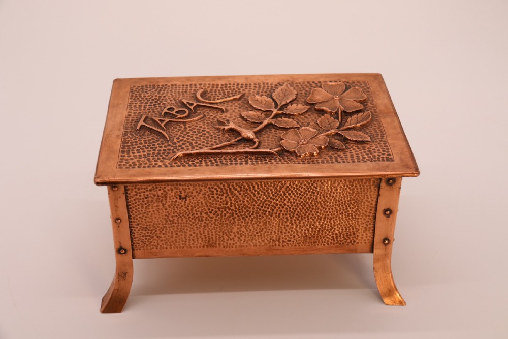 A rectangular Arts and Crafts copper box featuring a hand-hammered texture, splayed legs, and an embossed lid with a floral design and the word TABAC.