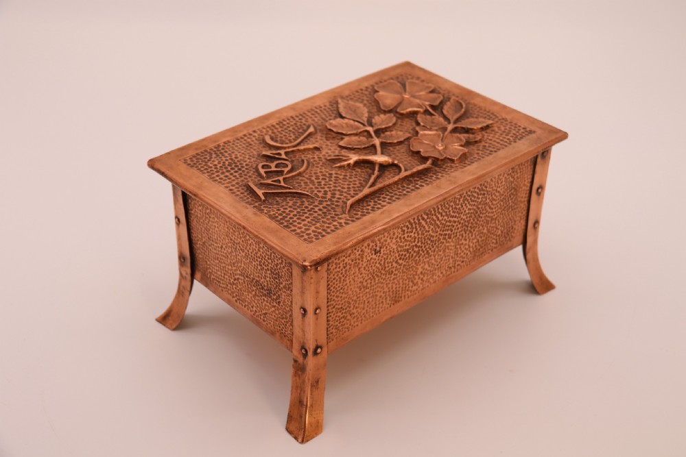 A rectangular Arts and Crafts copper box featuring a hand-hammered texture, splayed legs, and an embossed lid with a floral design and the word TABAC. View II