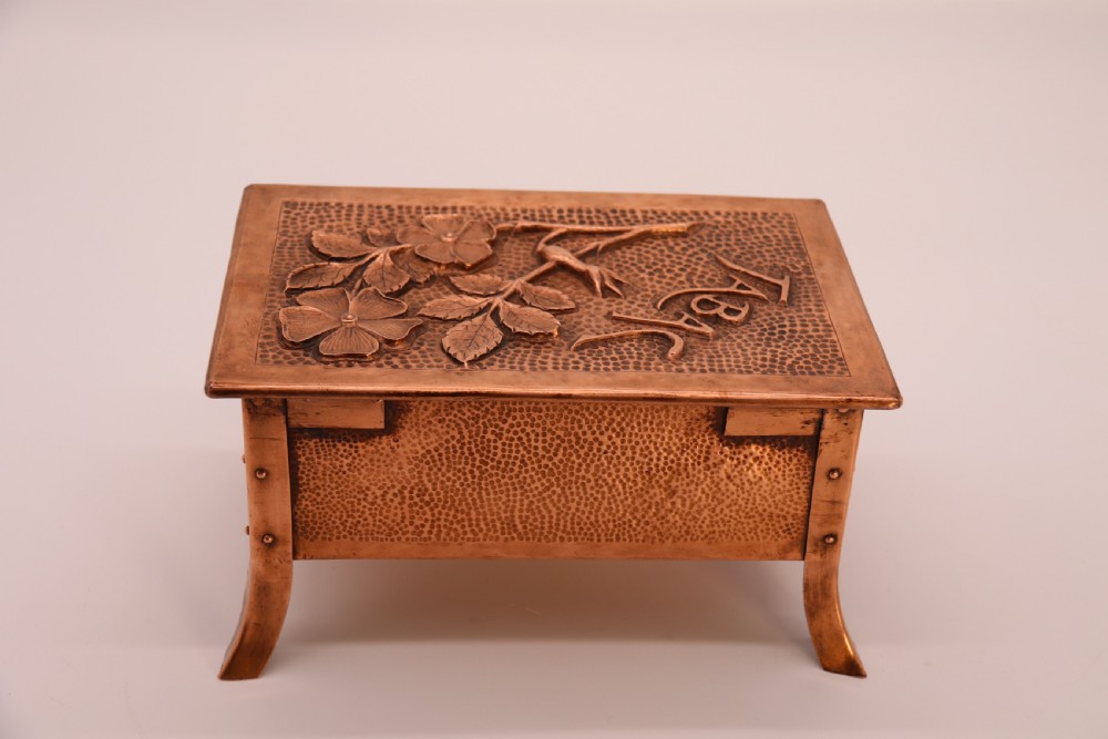 A rectangular Arts and Crafts copper box featuring a hand-hammered texture, splayed legs, and an embossed lid with a floral design and the word TABAC. View IV