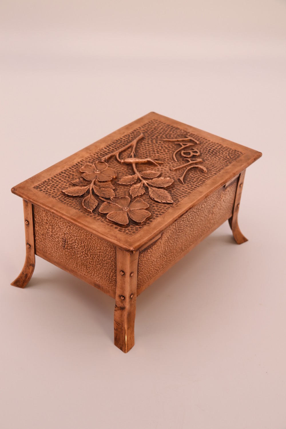 A rectangular Arts and Crafts copper box featuring a hand-hammered texture, splayed legs, and an embossed lid with a floral design and the word TABAC. View V