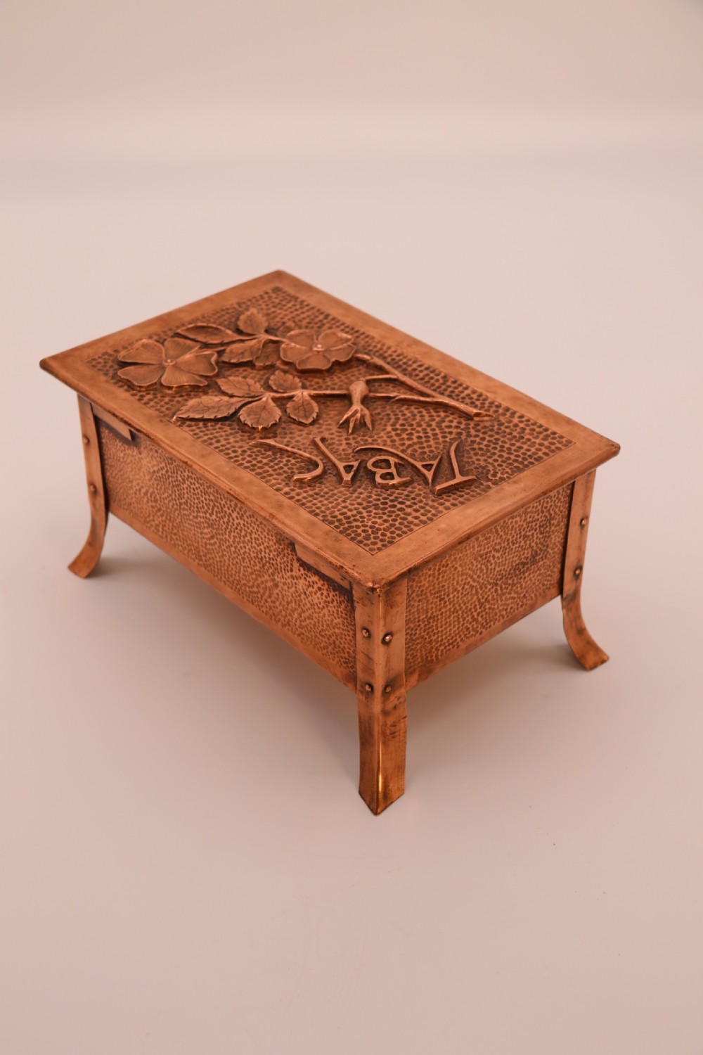 A rectangular Arts and Crafts copper box featuring a hand-hammered texture, splayed legs, and an embossed lid with a floral design and the word TABAC. View VI