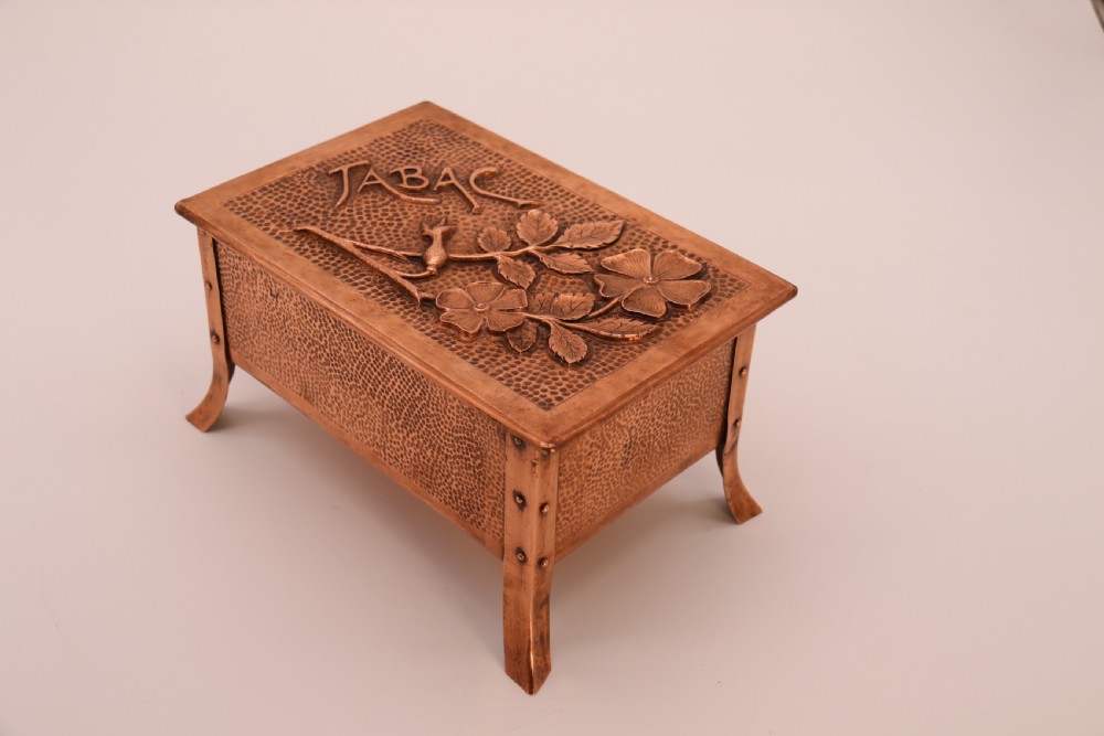 A rectangular Arts and Crafts copper box featuring a hand-hammered texture, splayed legs, and an embossed lid with a floral design and the word TABAC. View VIII