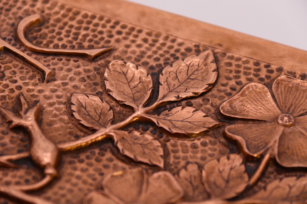 A rectangular Arts and Crafts copper box featuring a hand-hammered texture, splayed legs, and an embossed lid with a floral design and the word TABAC. View IX