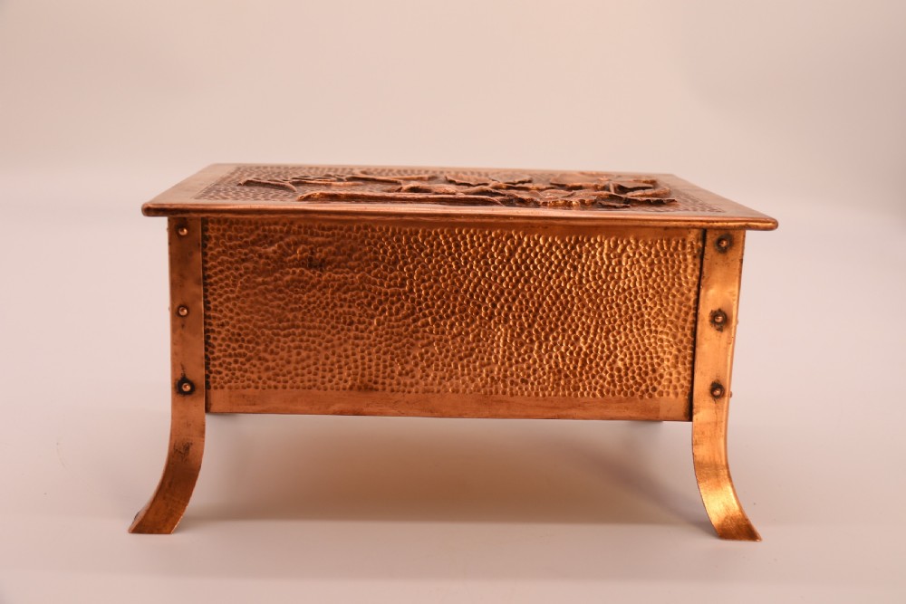 A rectangular Arts and Crafts copper box featuring a hand-hammered texture, splayed legs, and an embossed lid with a floral design and the word TABAC. View X