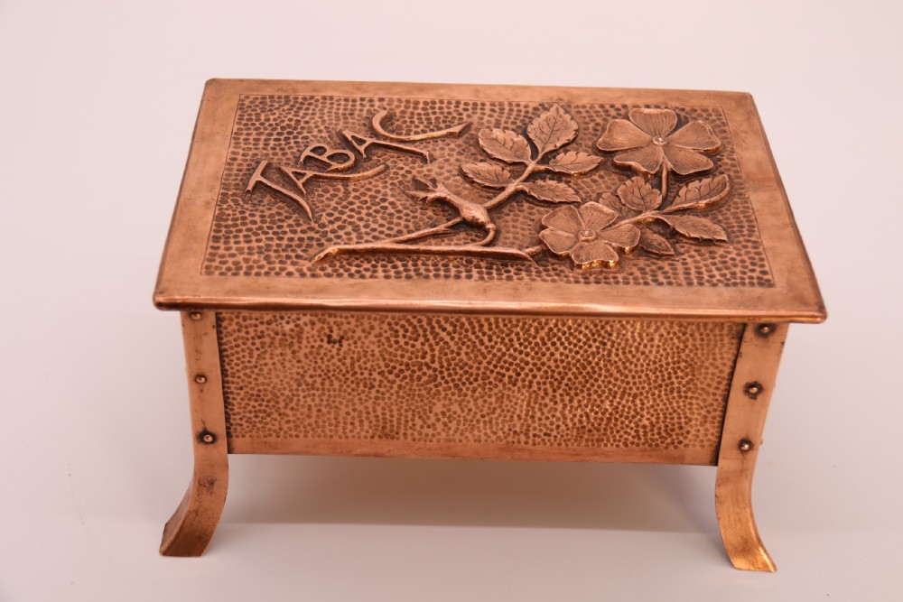 A rectangular Arts and Crafts copper box featuring a hand-hammered texture, splayed legs, and an embossed lid with a floral design and the word TABAC. View XV