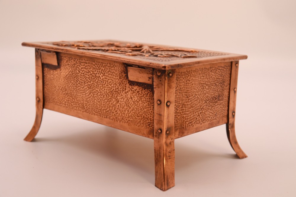 A rectangular Arts and Crafts copper box featuring a hand-hammered texture, splayed legs, and an embossed lid with a floral design and the word TABAC. View XVI