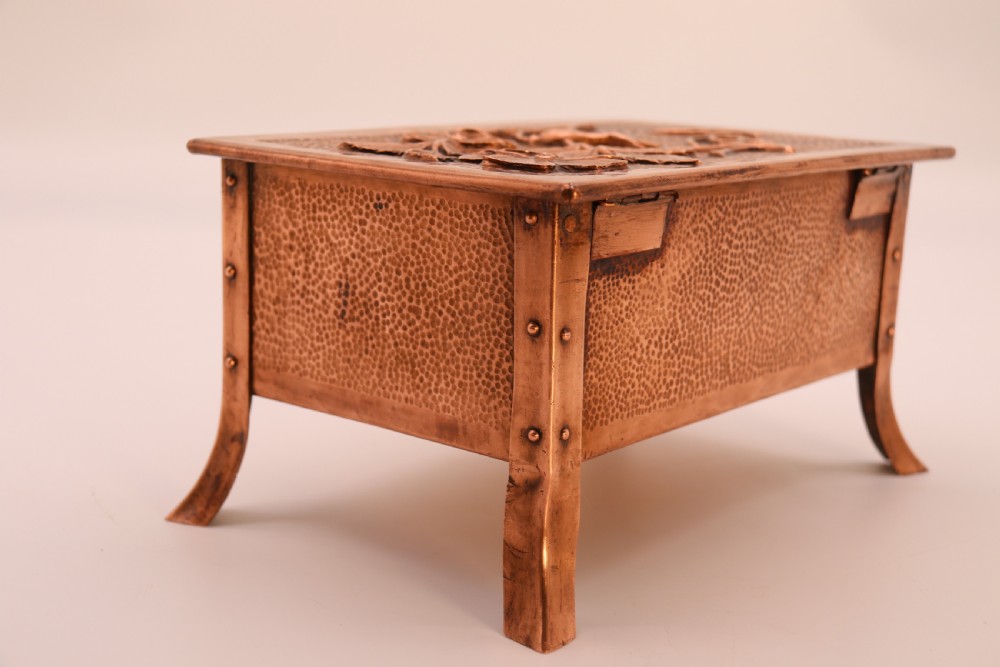 A rectangular Arts and Crafts copper box featuring a hand-hammered texture, splayed legs, and an embossed lid with a floral design and the word TABAC. View XX