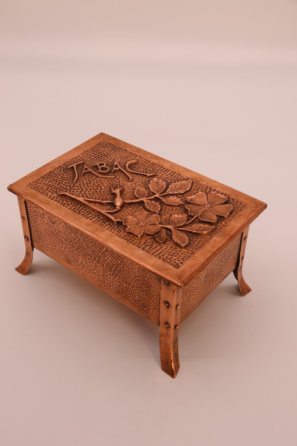 A rectangular Arts and Crafts copper box featuring a hand-hammered texture, splayed legs, and an embossed lid with a floral design and the word TABAC. View XXIV