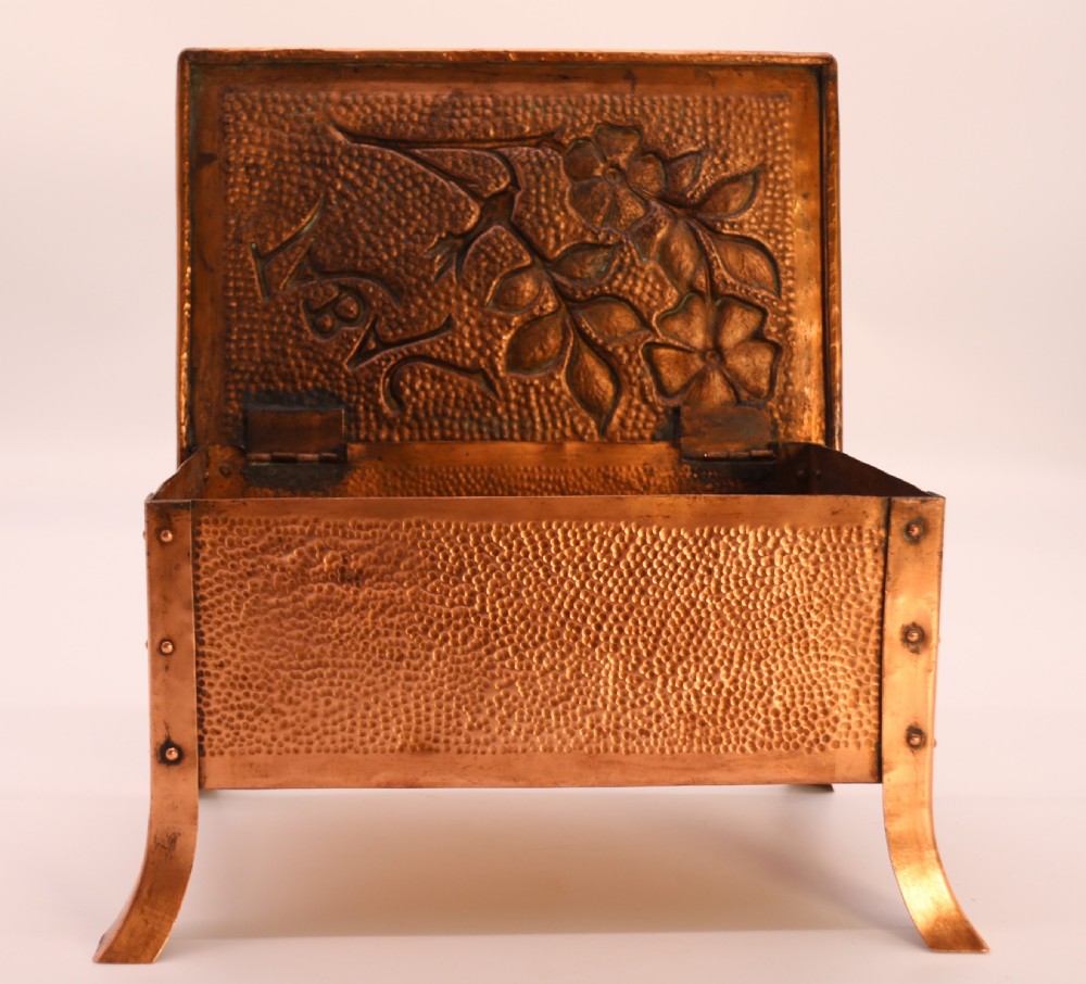 A rectangular Arts and Crafts copper box featuring a hand-hammered texture, splayed legs, and an embossed lid with a floral design and the word TABAC. View XXV