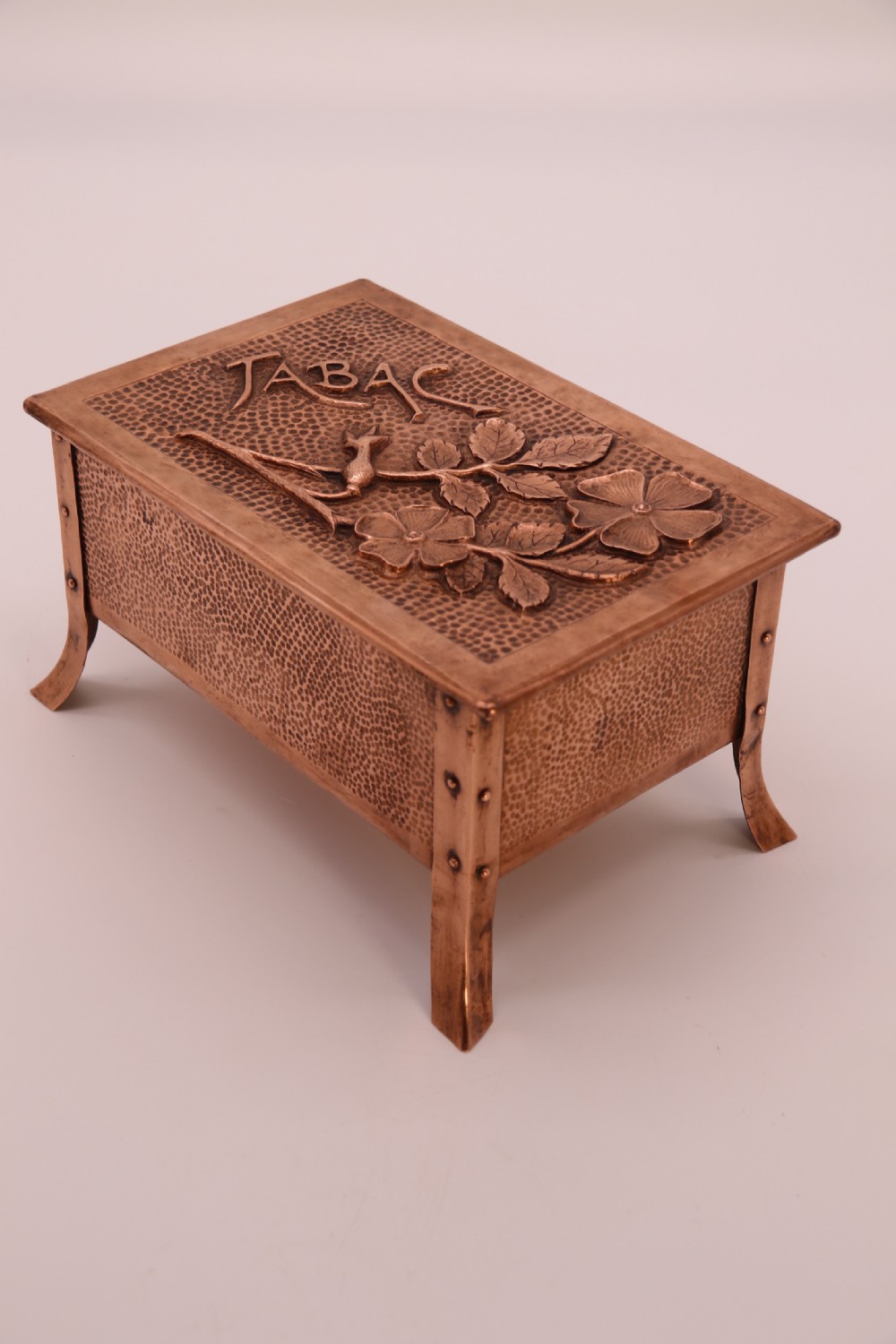 A rectangular Arts and Crafts copper box featuring a hand-hammered texture, splayed legs, and an embossed lid with a floral design and the word TABAC. View XXVI