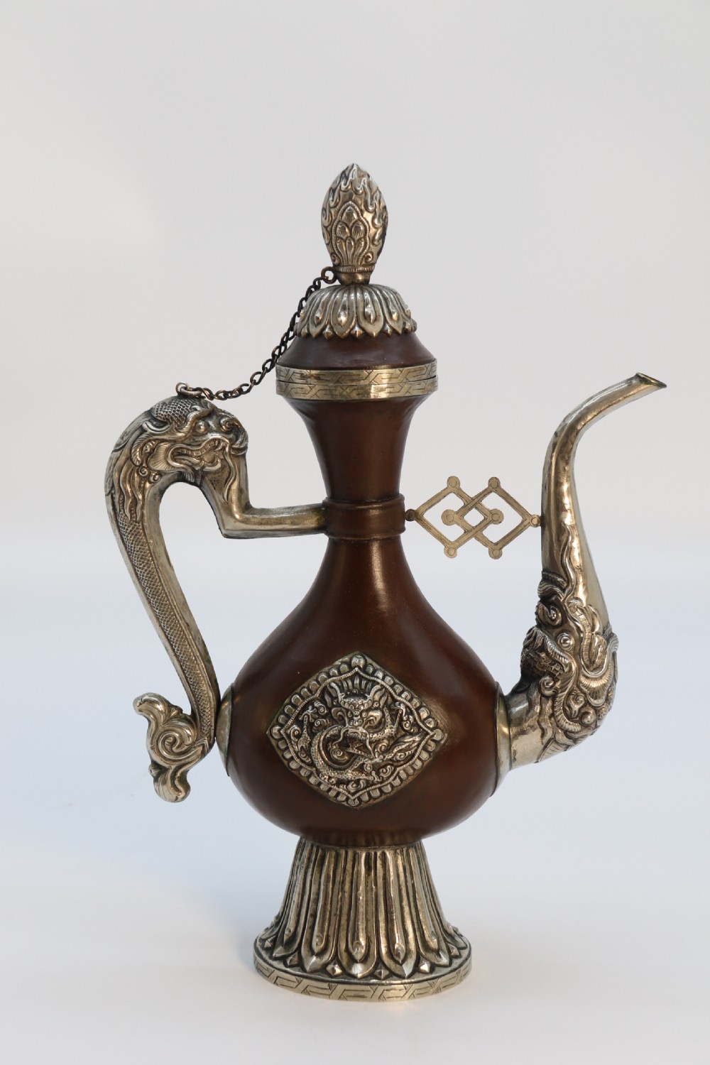 An antique Indo-Tibetan ritual ewer featuring a dark brown bulbous body, repousse silver dragon spout and handle, lotus base, and chained lid finial.
