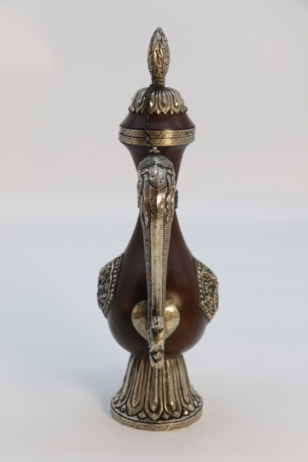 An antique Indo-Tibetan ritual ewer featuring a dark brown bulbous body, repousse silver dragon spout and handle, lotus base, and chained lid finial. View V