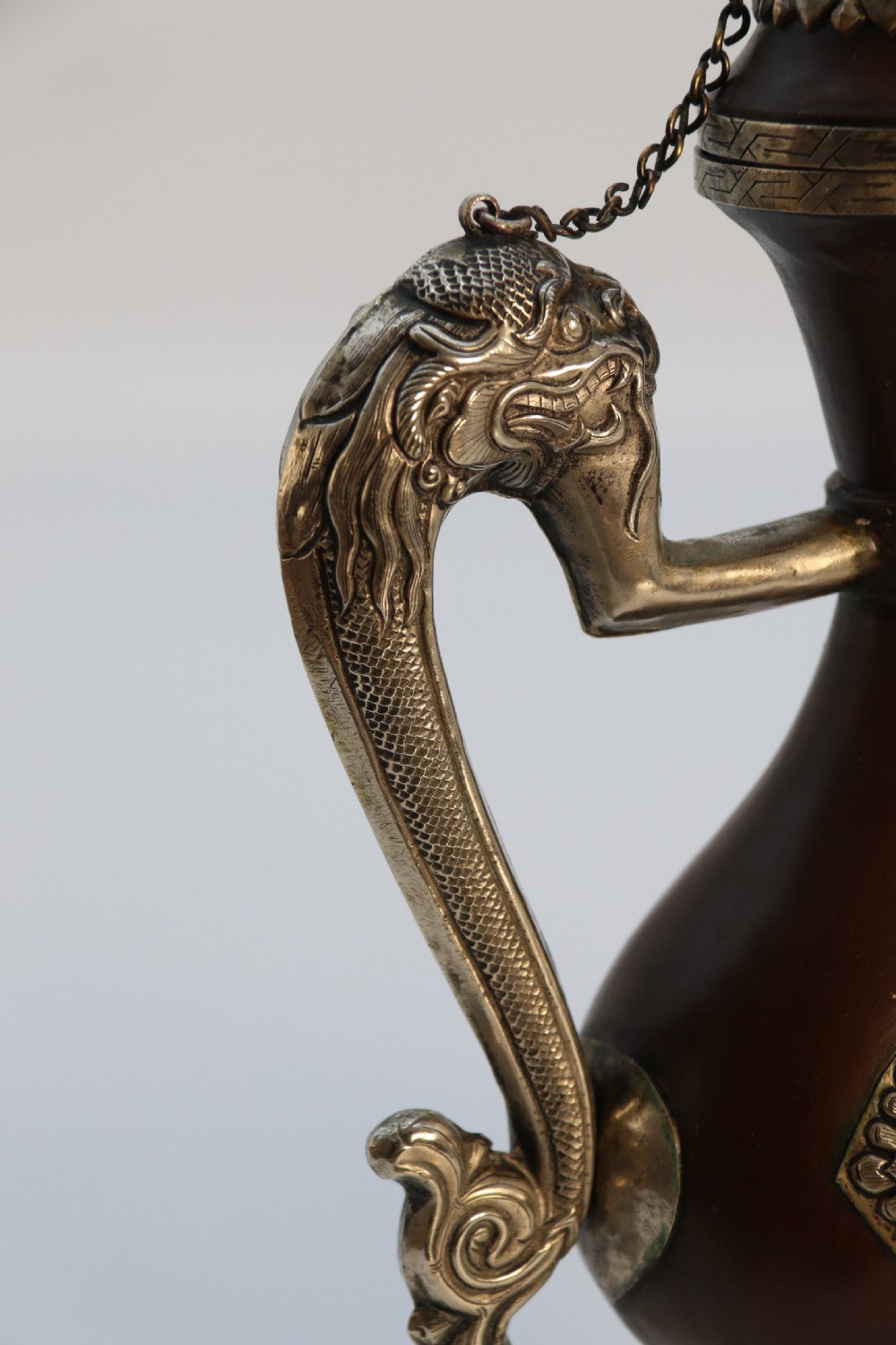 An antique Indo-Tibetan ritual ewer featuring a dark brown bulbous body, repousse silver dragon spout and handle, lotus base, and chained lid finial. View XIV
