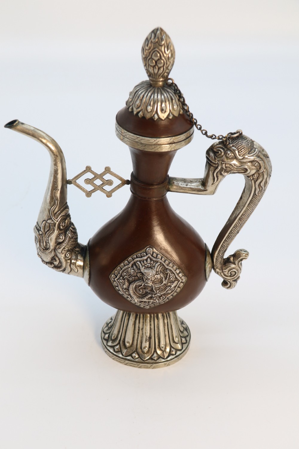 An antique Indo-Tibetan ritual ewer featuring a dark brown bulbous body, repousse silver dragon spout and handle, lotus base, and chained lid finial. View XVI