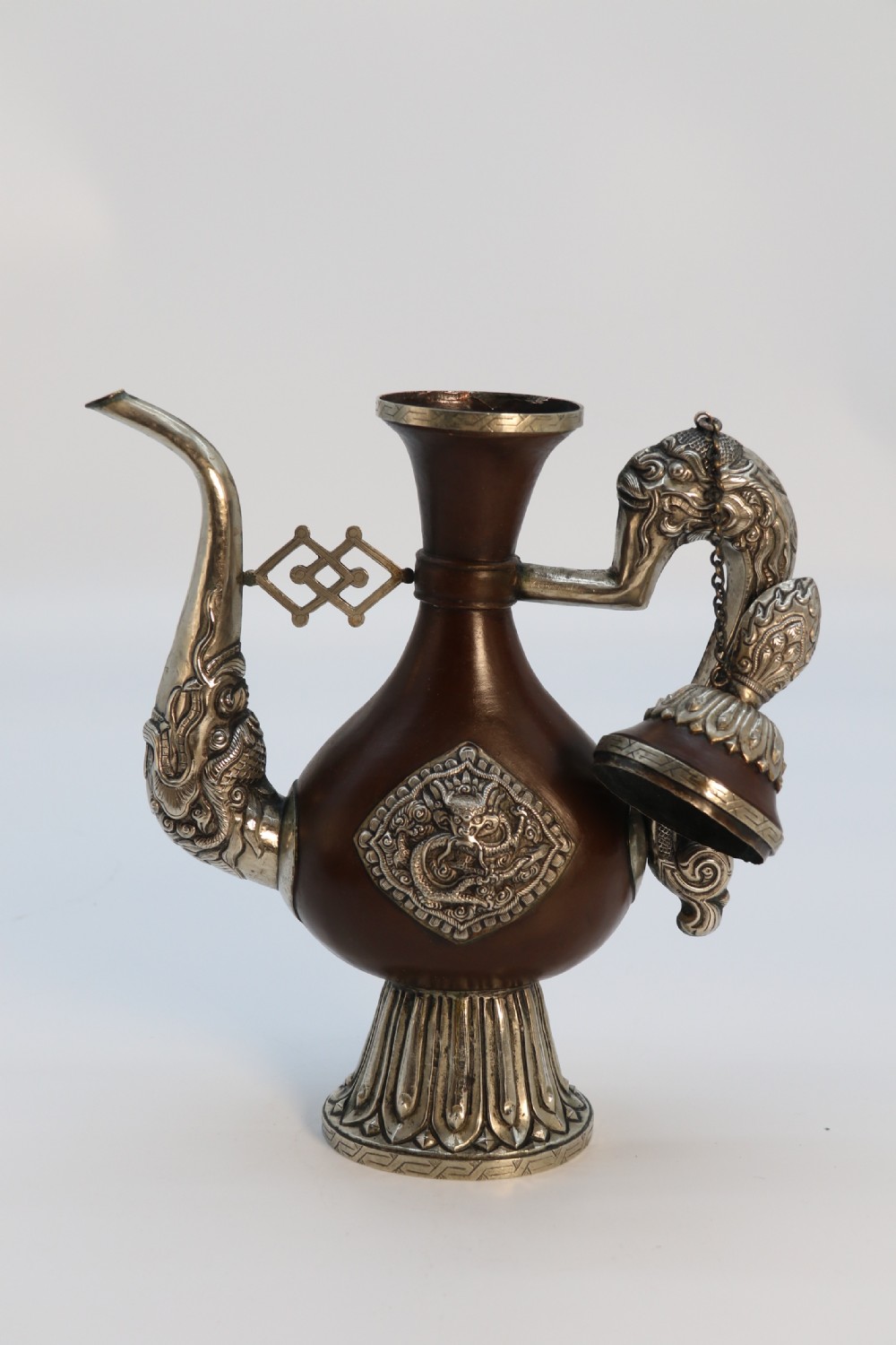 An antique Indo-Tibetan ritual ewer featuring a dark brown bulbous body, repousse silver dragon spout and handle, lotus base, and chained lid finial. View XVII