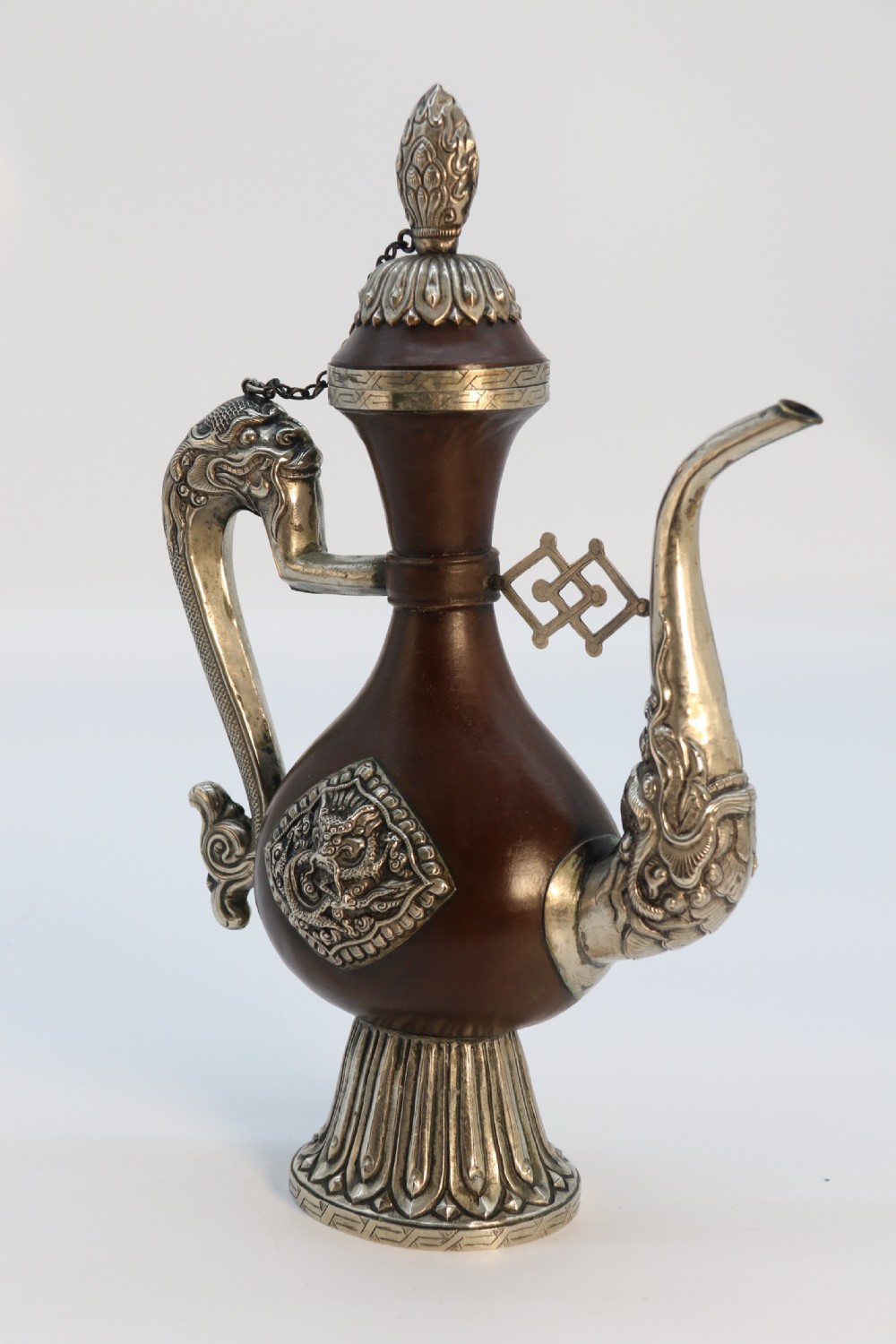 An antique Indo-Tibetan ritual ewer featuring a dark brown bulbous body, repousse silver dragon spout and handle, lotus base, and chained lid finial. View XX