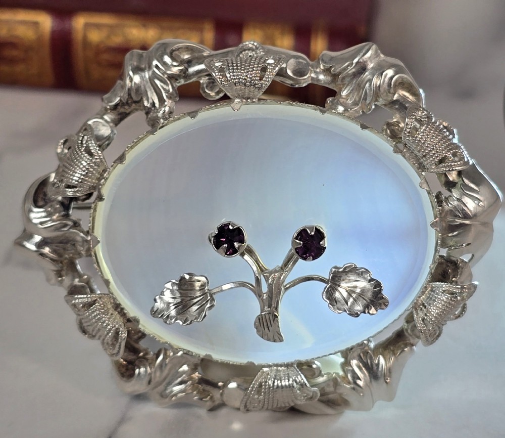 An oval translucent chalcedony brooch set within a foliate silver frame, featuring a silver botanical spray with two purple amethyst stones at the centre. View III