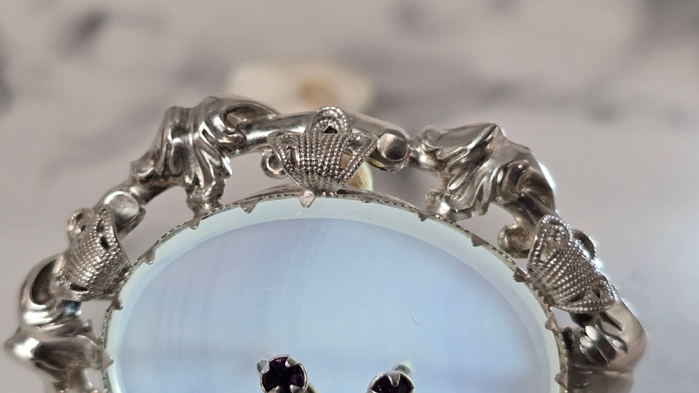 An oval translucent chalcedony brooch set within a foliate silver frame, featuring a silver botanical spray with two purple amethyst stones at the centre. View V