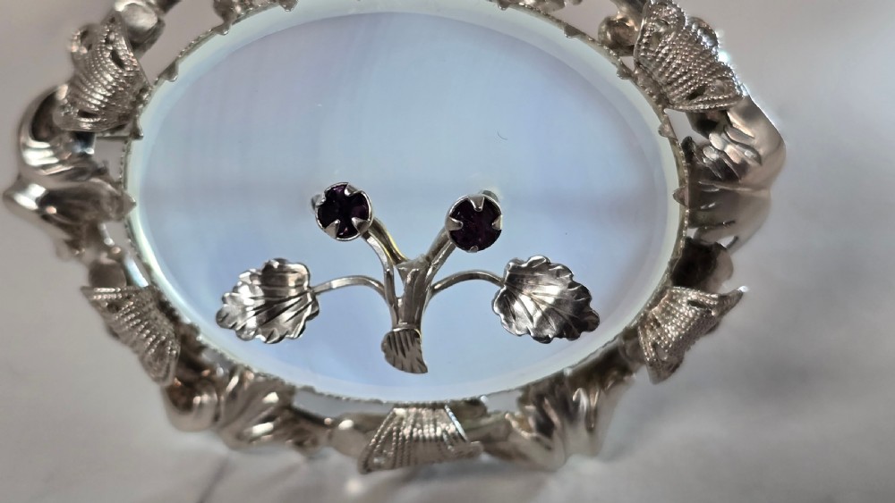 An oval translucent chalcedony brooch set within a foliate silver frame, featuring a silver botanical spray with two purple amethyst stones at the centre. View VI