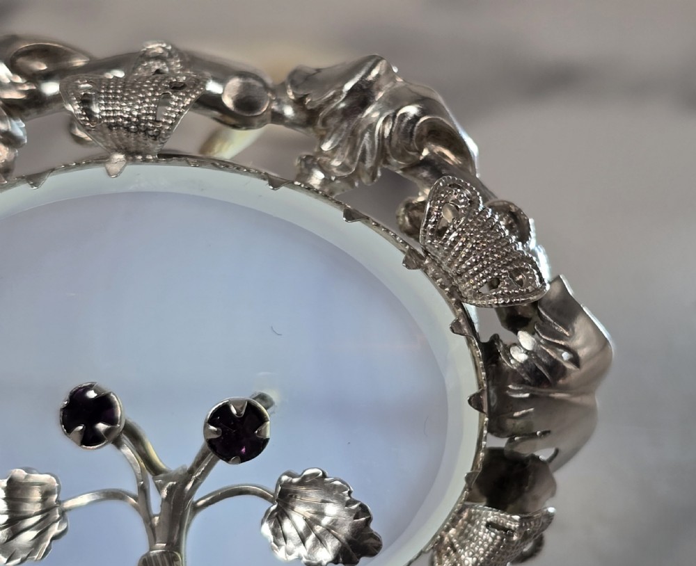 An oval translucent chalcedony brooch set within a foliate silver frame, featuring a silver botanical spray with two purple amethyst stones at the centre. View VII