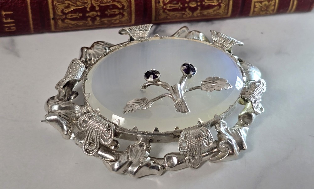An oval translucent chalcedony brooch set within a foliate silver frame, featuring a silver botanical spray with two purple amethyst stones at the centre. View VIII