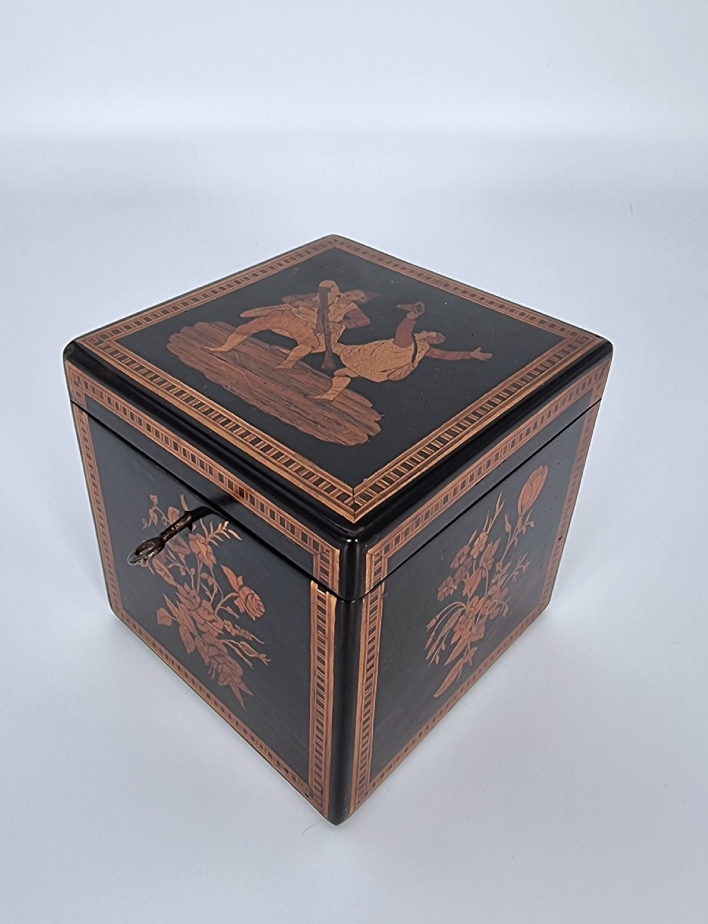 A square ebonised wooden box with Sorrento marquetry inlay depicting figures on the lid, floral sprays on the sides, and a key inserted in the keyhole.
