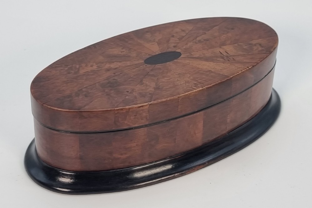 An oval wooden box constructed with radially segmented burr walnut veneer, a dark central oval inlay, and a moulded ebonised base.