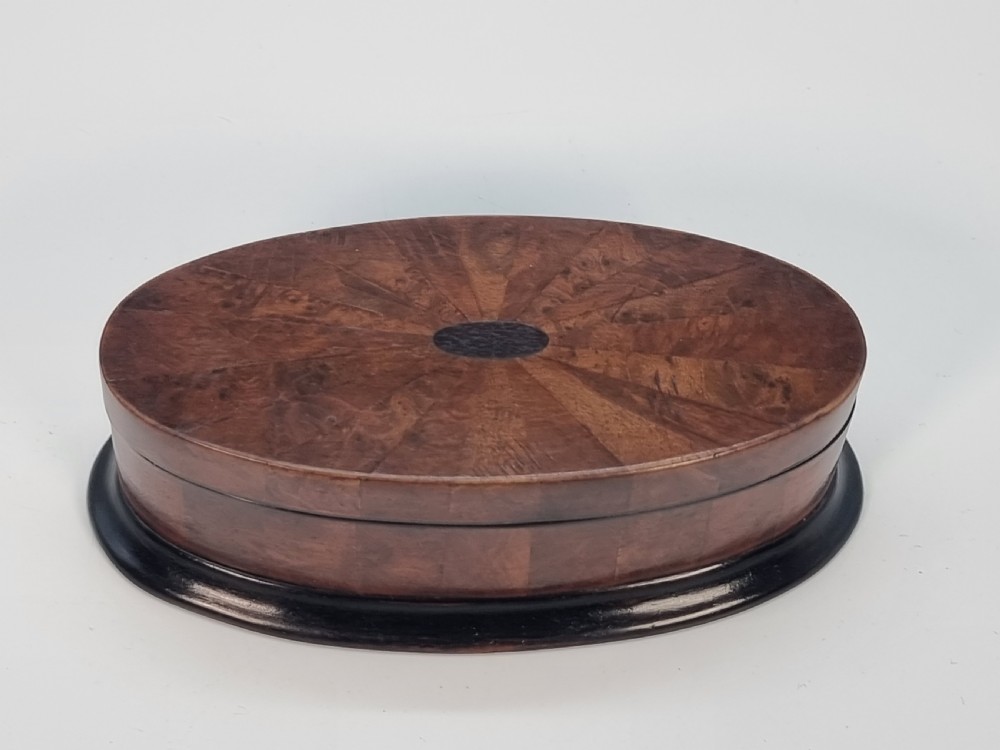 An oval wooden box constructed with radially segmented burr walnut veneer, a dark central oval inlay, and a moulded ebonised base. View III