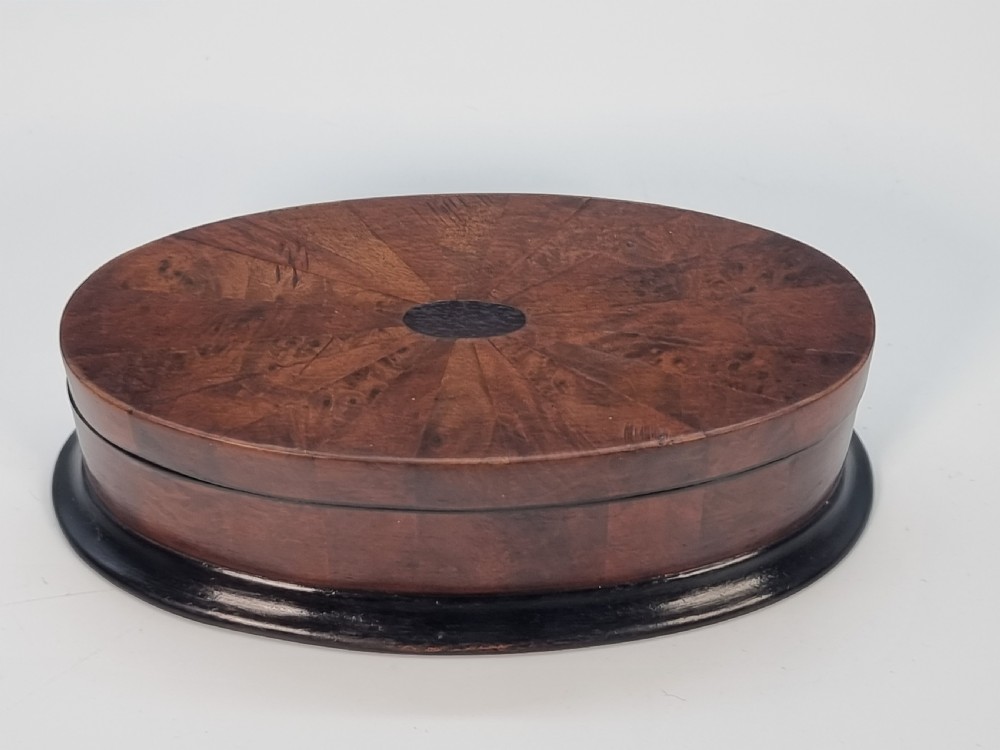 An oval wooden box constructed with radially segmented burr walnut veneer, a dark central oval inlay, and a moulded ebonised base. View VI