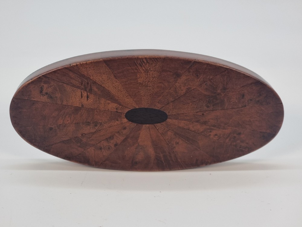 An oval wooden box constructed with radially segmented burr walnut veneer, a dark central oval inlay, and a moulded ebonised base. View VIII