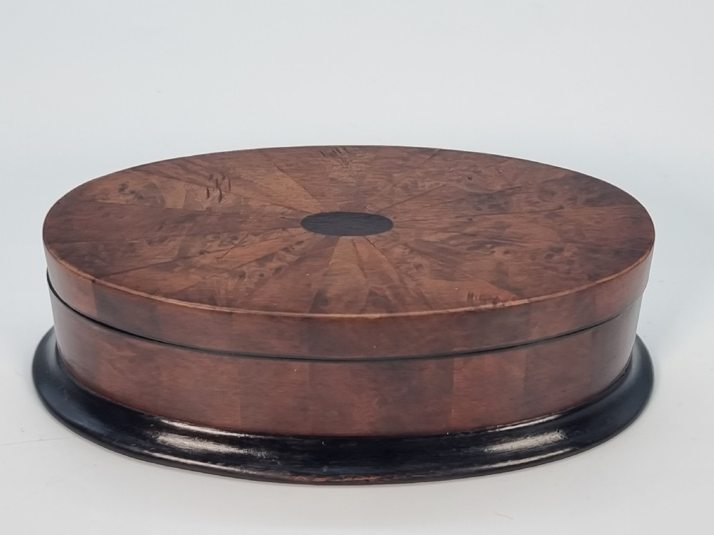 An oval wooden box constructed with radially segmented burr walnut veneer, a dark central oval inlay, and a moulded ebonised base. View X