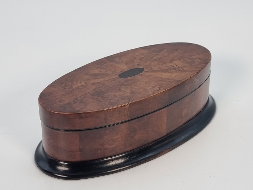 An oval wooden box constructed with radially segmented burr walnut veneer, a dark central oval inlay, and a moulded ebonised base. View XI