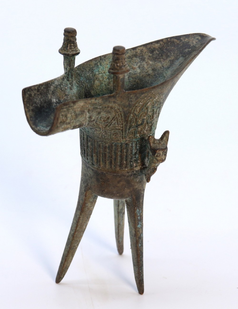 An archaic Chinese patinated bronze tripod wine vessel featuring flared splayed legs, a extended pouring spout with two capped upright posts, and carved relief geometric bands.