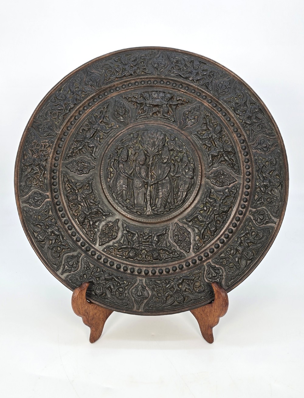 A circular dark metal charger featuring intricate repousse depicting central figures under a tree surrounded by concentric decorative borders.