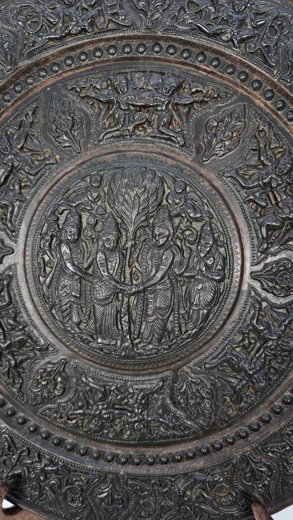 A circular dark metal charger featuring intricate repousse depicting central figures under a tree surrounded by concentric decorative borders. View VIII