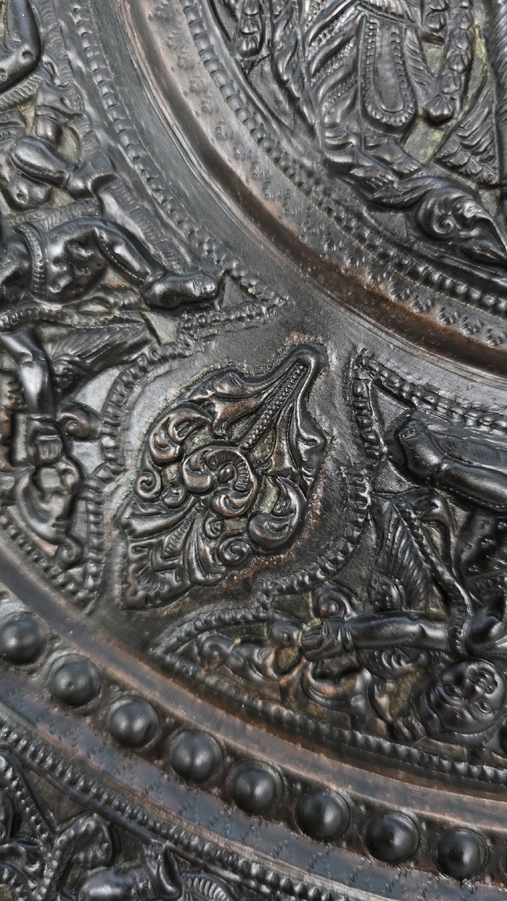 A circular dark metal charger featuring intricate repousse depicting central figures under a tree surrounded by concentric decorative borders. View IX
