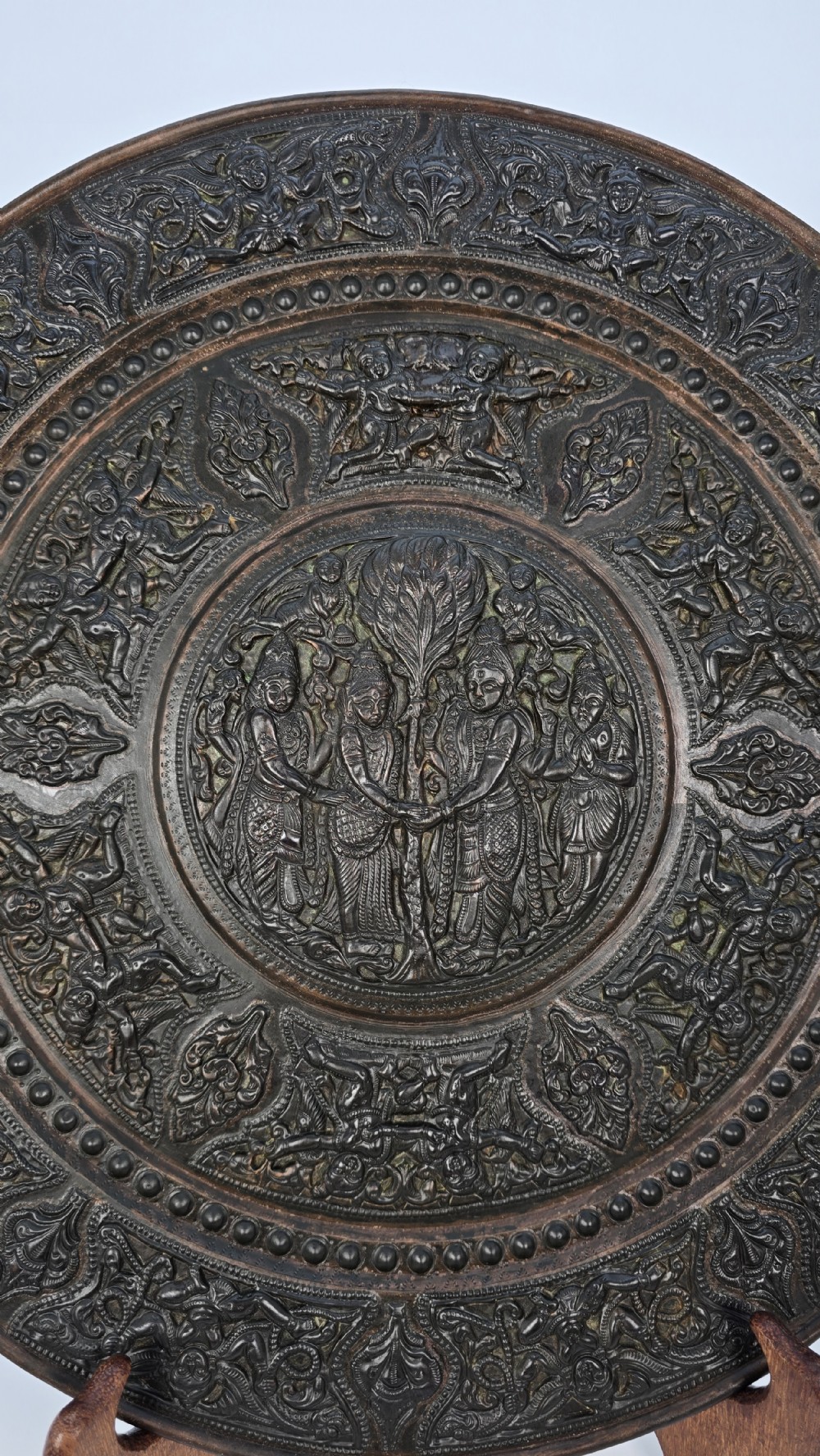 A circular dark metal charger featuring intricate repousse depicting central figures under a tree surrounded by concentric decorative borders. View X