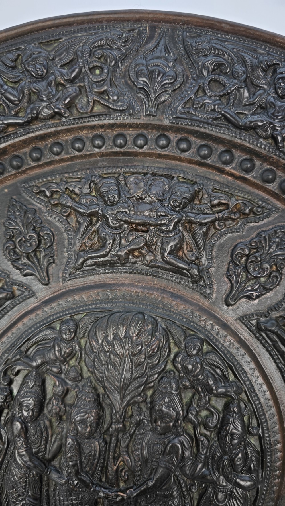 A circular dark metal charger featuring intricate repousse depicting central figures under a tree surrounded by concentric decorative borders. View XI