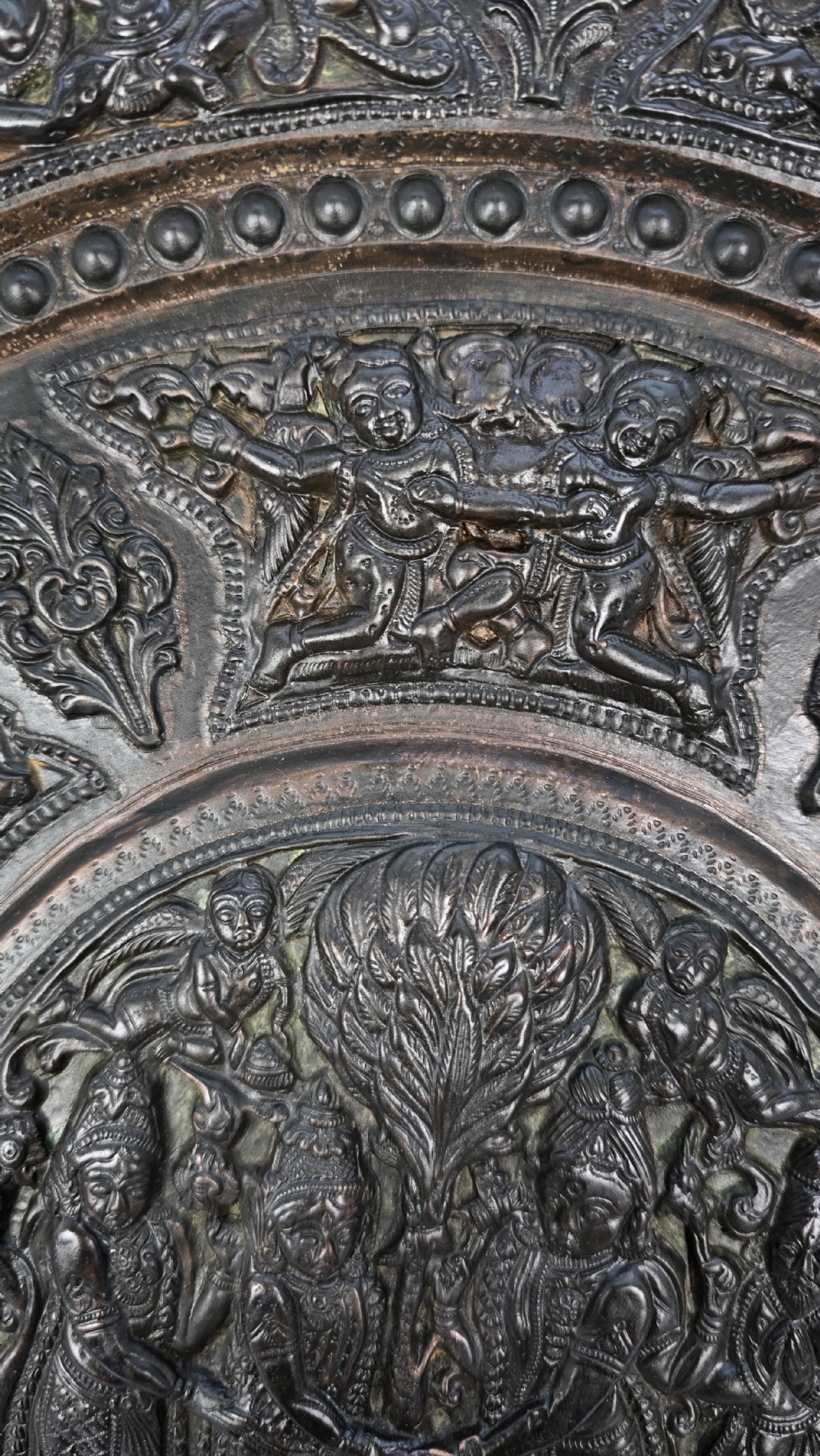 A circular dark metal charger featuring intricate repousse depicting central figures under a tree surrounded by concentric decorative borders. View XII