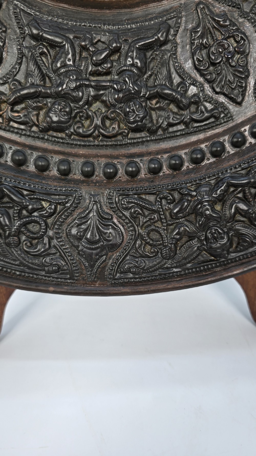 A circular dark metal charger featuring intricate repousse depicting central figures under a tree surrounded by concentric decorative borders. View XIV