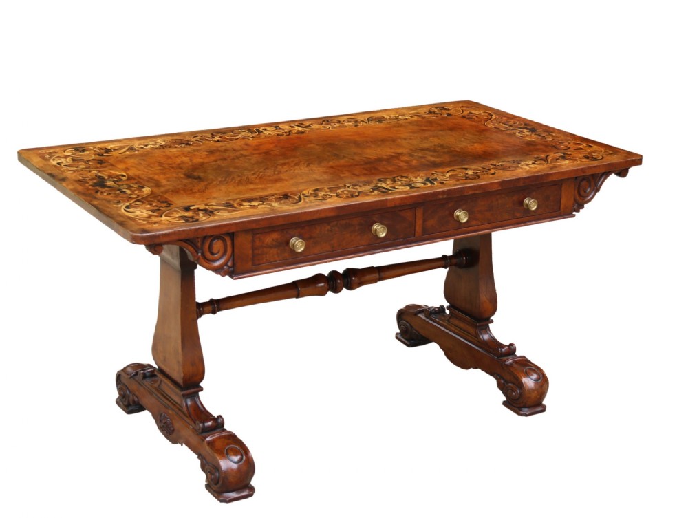 An early 19th-century flame mahogany library table featuring floral marquetry top borders, two frieze drawers with brass knobs, and carved trestle legs connected by a turned stretcher.