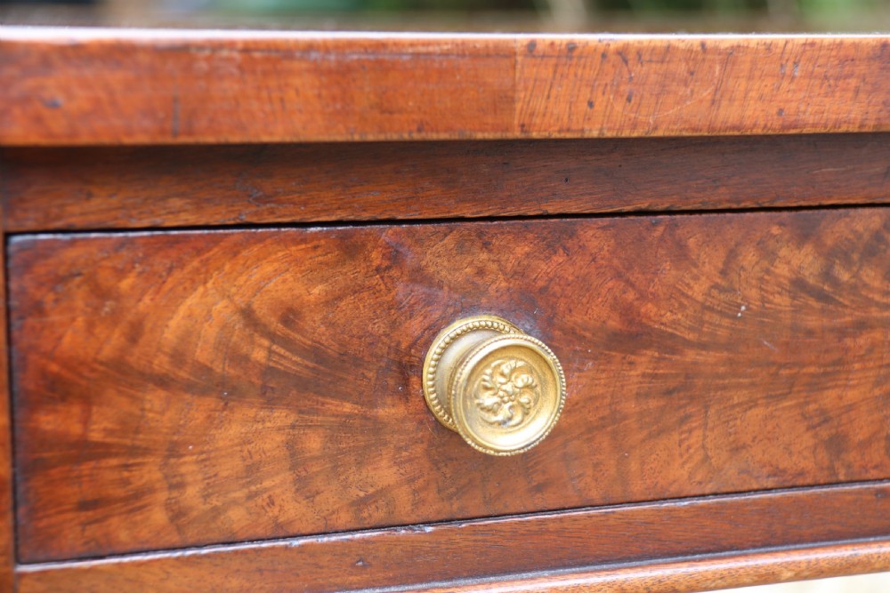 An early 19th-century flame mahogany library table featuring floral marquetry top borders, two frieze drawers with brass knobs, and carved trestle legs connected by a turned stretcher. View XVI